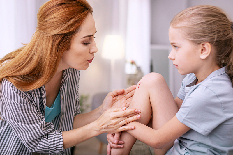Ein Kind mit einer sichtbaren Schürfwunde auf dem Knie, die sorgfältig mit Bepanthen Wund- und Heilsalbe behandelt wird, um die Heilung zu fördern und Infektionen vorzubeugen. Das Kind wirkt beruhigt und erleichtert, während die Salbe sanft auf die Wunde aufgetragen wird.